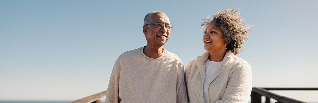 Mature couple smiling happily while taking a walk along a wooden foot bridge at the beach.